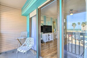 Une pièce dotée d’une porte-fenêtre coulissante donnant sur un balcon avec vue sur des palmiers. À l’intérieur, on trouve un meuble blanc avec un téléviseur et une chaise blanche.