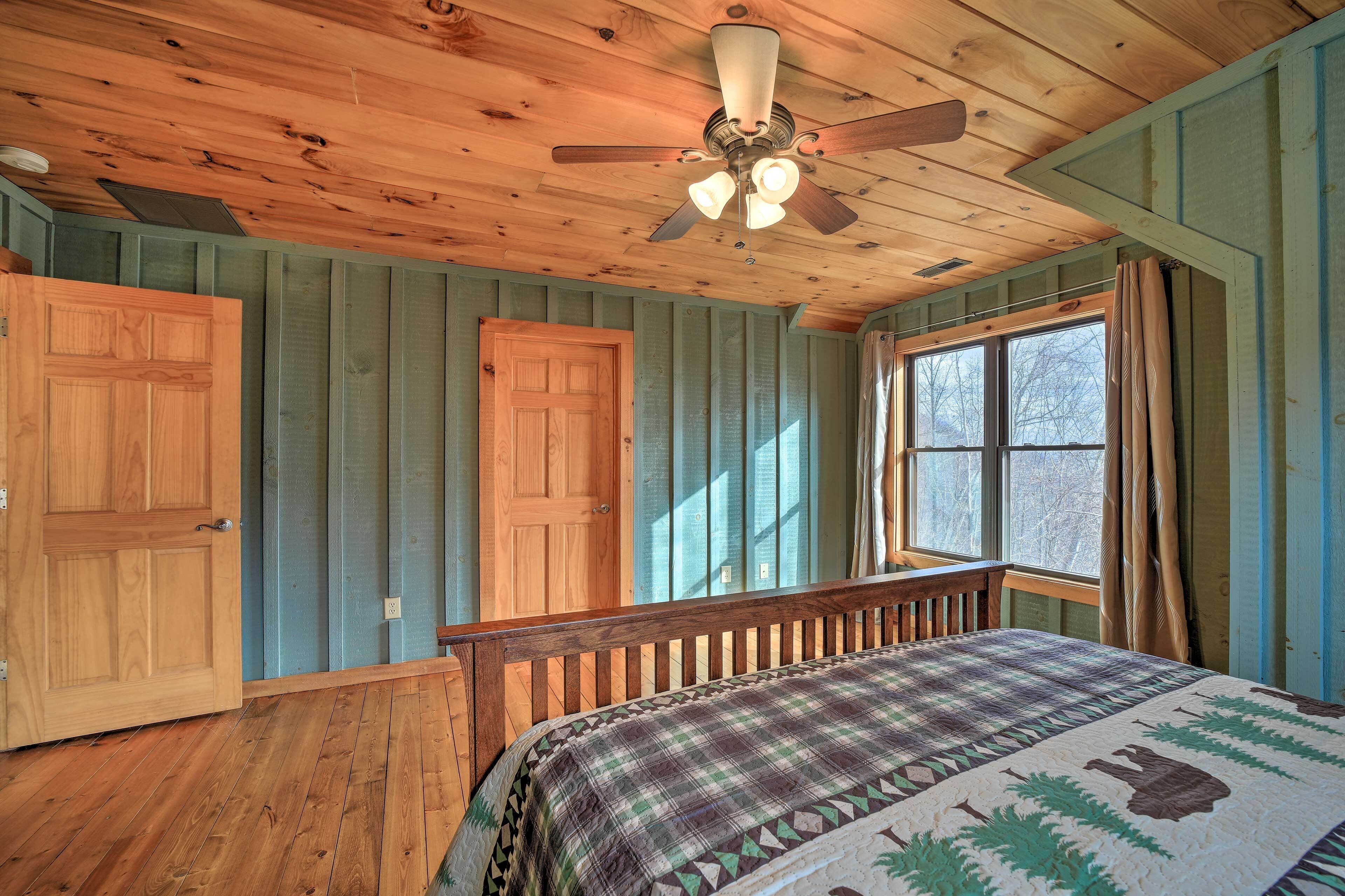 A bedroom with a wooden ceiling, a bed with a plaid bedspread, a window with curtains, and a door.