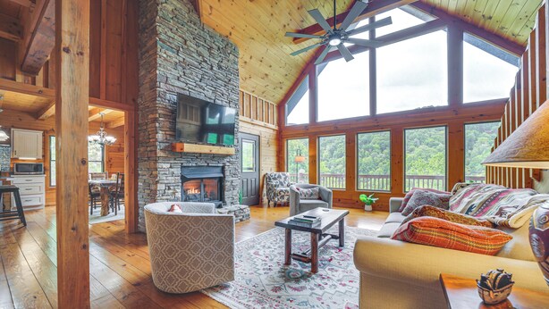 A spacious living room with a stone fireplace, wooden ceiling beams, and large windows offering a view of the outdoors. - Decks & Mountain Views: Waynesville Getaway! (Waynesville)