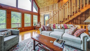 A spacious living room with a wooden staircase, large windows, and a cozy sofa with patterned cushions. - Decks & Mountain Views: Waynesville Getaway! (Waynesville)