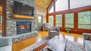 A cozy living room with a stone fireplace, wooden floors, and large windows offering a view of the outdoors. - Decks & Mountain Views: Waynesville Getaway! (Waynesville)