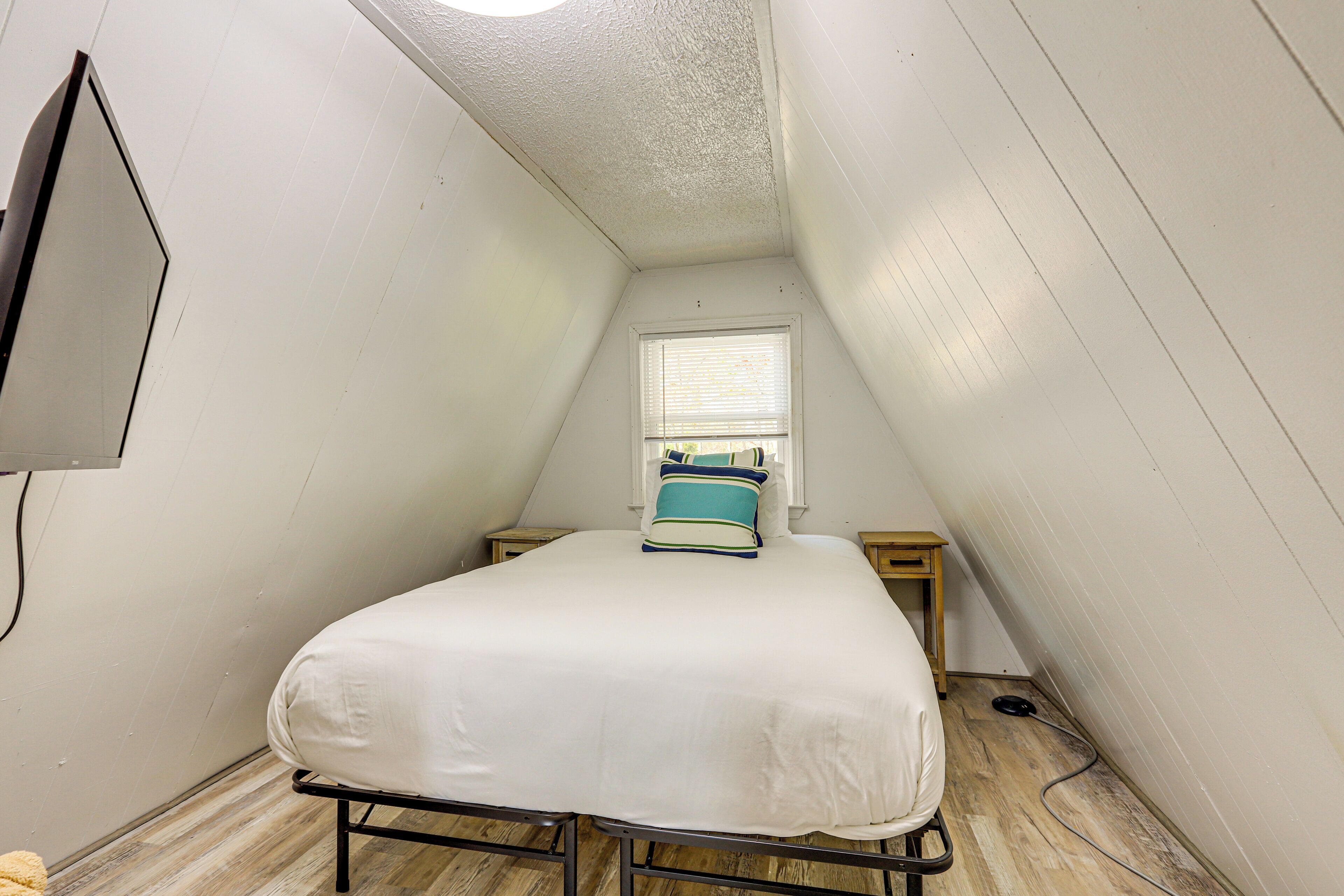 A small, triangular attic bedroom with a single bed, a window with blinds, and a wall-mounted TV.