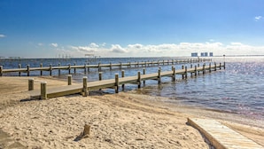 A beach with a wooden pier extending into calm waters, a sandy shore, and distant buildings.