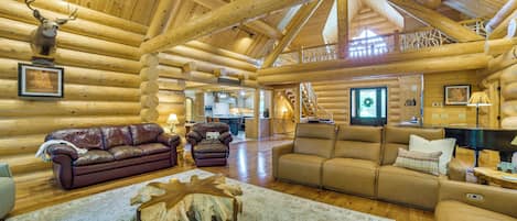 A log cabin interior with a fireplace, leather sofas, a dining area, and a staircase leading to an upper level.