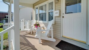 Un porche con sillones Adirondack blancos, una mesita con una planta en maceta y una puerta con ventanal.
