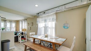 Un comedor con una mesa de madera, sillas blancas y un banco. Se ve una ventana con cortinas y un cartel en la pared.