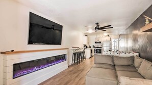 Een moderne woonkamer met een open haard, een bank, een aan de muur bevestigde televisie en een keuken op de achtergrond.