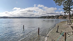 A pebbled beach with clear water, a distant shoreline, and a tree on the right. - Gorgeous Poulsbo Waterfront Home on Liberty Bay! (Poulsbo)