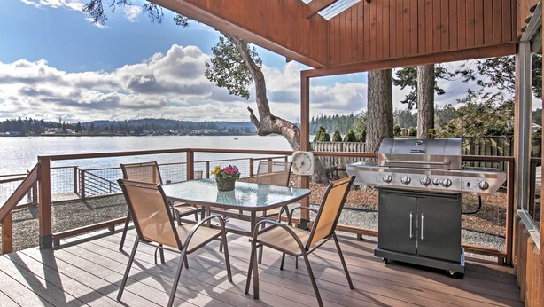 A wooden deck with a glass table and chairs, a grill, and a scenic view of a lake and mountains. - Gorgeous Poulsbo Waterfront Home on Liberty Bay! (Poulsbo)