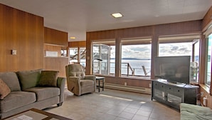 A living room with a sofa, armchair, and television, large windows with a view of the water, and a fireplace. - Gorgeous Poulsbo Waterfront Home on Liberty Bay! (Poulsbo)