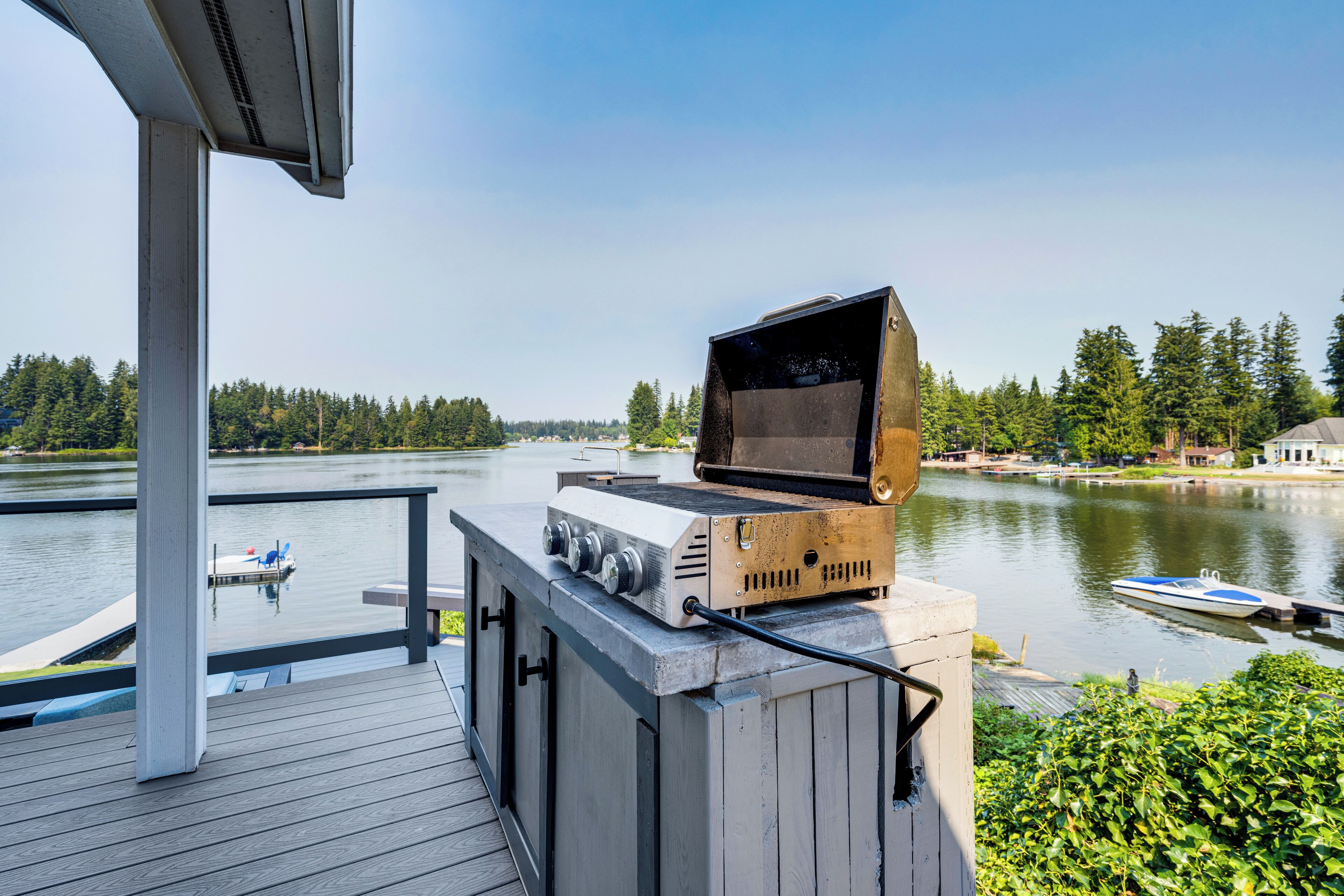 A barbecue grill on a wooden deck overlooking a lake with boats and trees in the background.