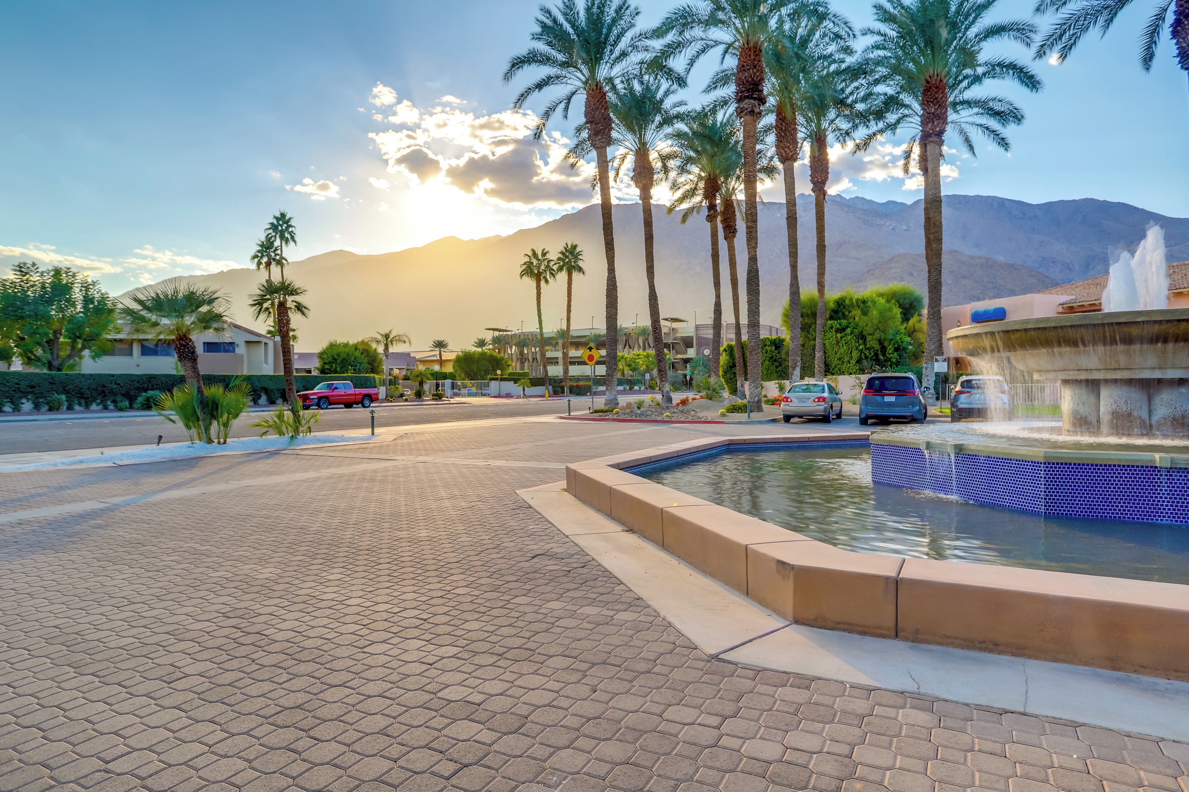A paved walkway with a water feature, palm trees, and a mountain in the background.