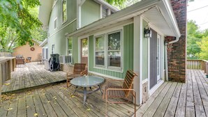 A wooden deck with a glass table and two chairs, a brick chimney, and a green building with multiple windows. - Pet-friendly Home w/ Views of Lake Delavan! (Delavan)