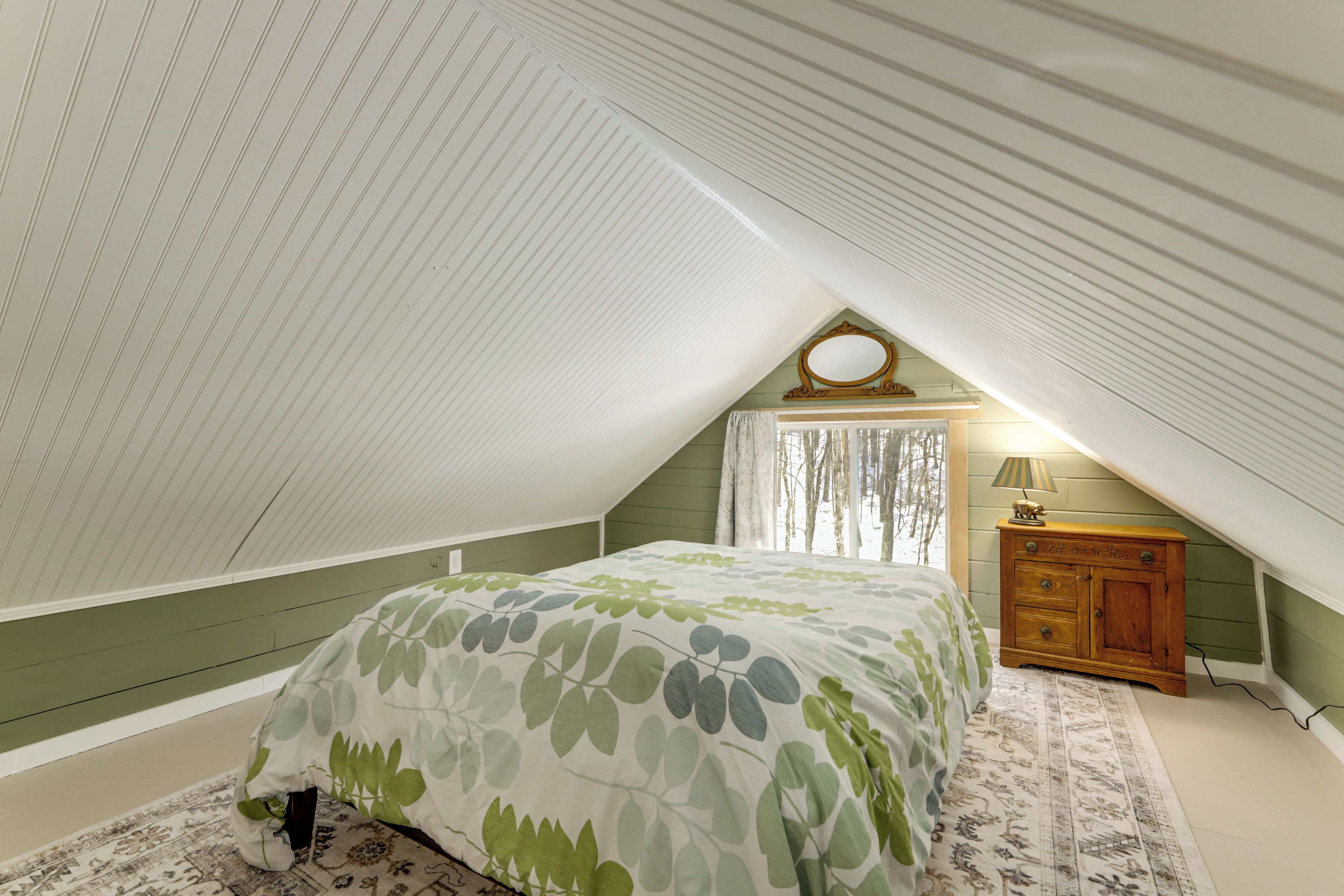Un dormitorio con techo inclinado, una cama con cubrecama floral, un aparador de madera y una ventana con cortinas.