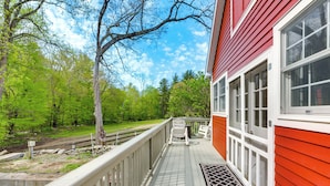 A wooden deck with a white railing, a red building with large windows, and a green landscape with trees. - Killingly Barnyard Studio in Wine Country! (Dayville)