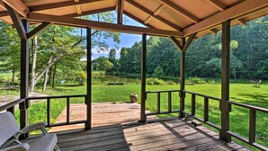 Une terrasse en bois couverte, dotée d’un banc, qui donne sur une zone herbeuse agrémentée d’arbres et d’un étang.