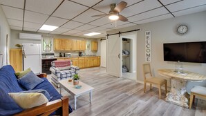Un vaste salon comprenant un canapé, une table basse et un coin repas avec une table et des chaises. Au fond, on aperçoit une cuisine avec des meubles en bois et un réfrigérateur.