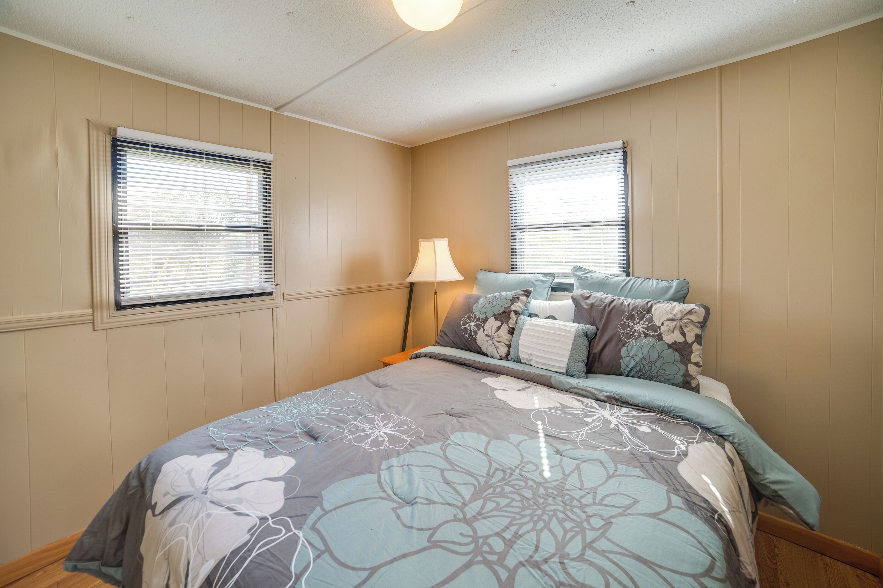 Un dormitorio con dos ventanas, una cama con ropa de cama estampada y una lámpara de noche.