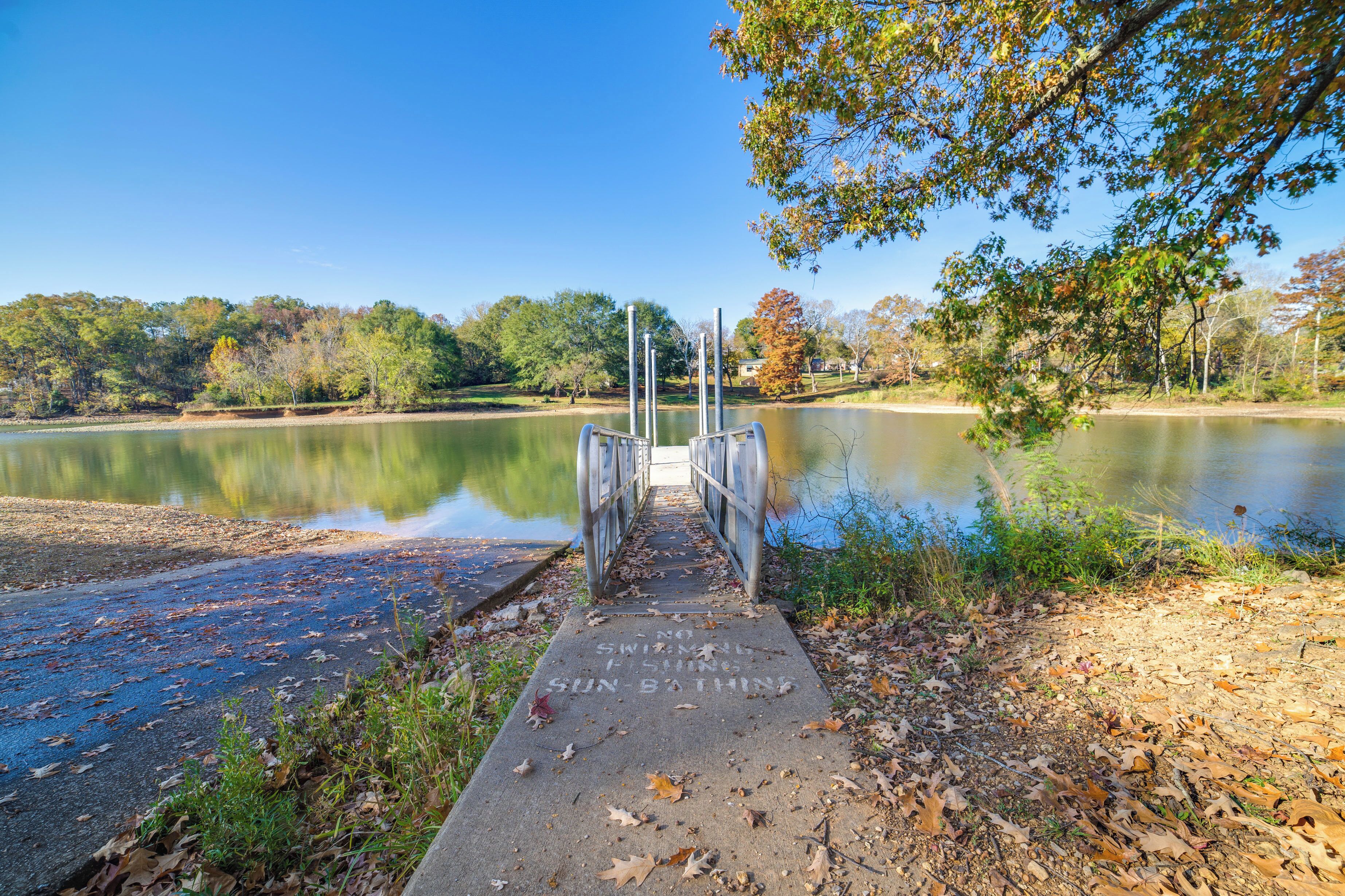 A concrete pathway leading to a metal bridge over a calm lake, surrounded by trees and a clear sky.