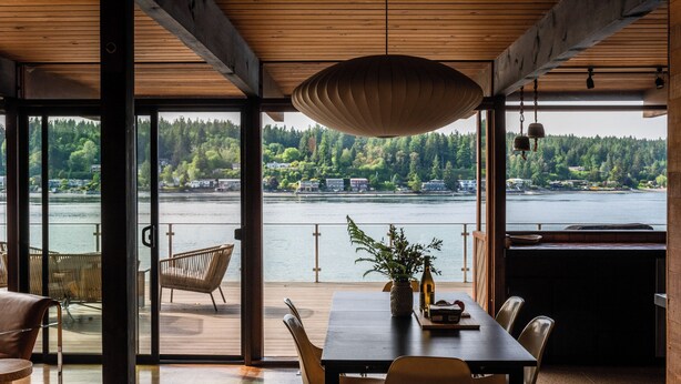 Comedor con vistas a un lago y un bosque, que presenta techo de madera, ventanas amplias y una lámpara colgante.
