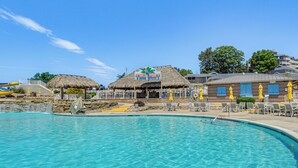 Un espace piscine avec un bâtiment à toit de chaume portant l’inscription « TIKI HUT » et un bâtiment en briques avec des volets bleus.