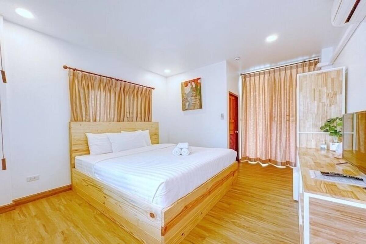 A bedroom with a wooden bed, white bedding, a desk with a plant, and curtains.