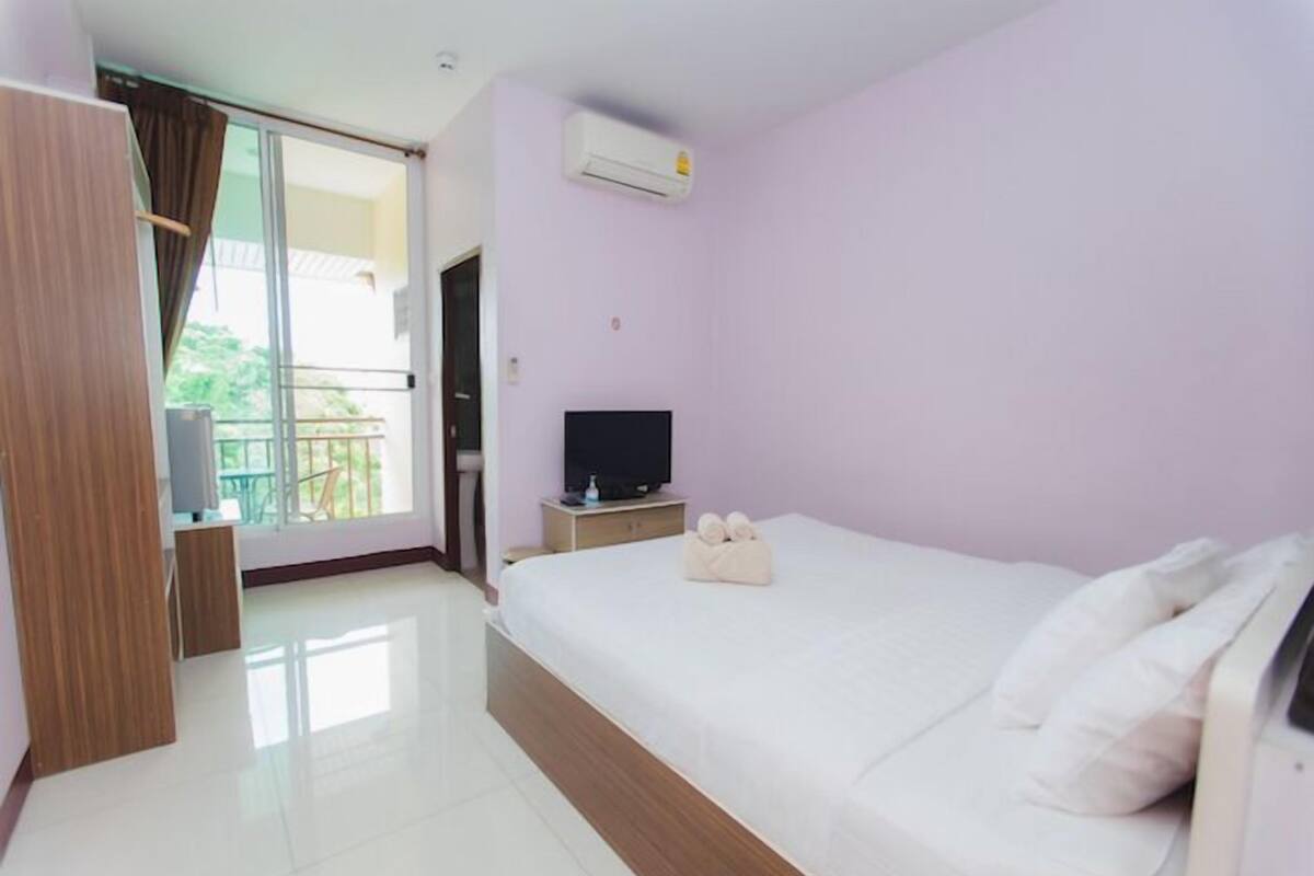 A hotel room with a large bed, a TV on a wooden nightstand, a balcony with a view, and an air conditioning unit on the wall.