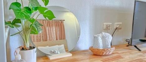 A room with a wooden floor, a large mirror, a potted plant, a bedside table with a bottle of hand sanitizer, a basket, and a bed with a wooden headboard.
