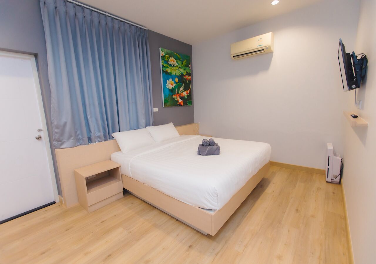 A modern bedroom with a wooden floor, a bed with white bedding, a small bedside table, a wall-mounted air conditioner, and a painting of flowers.
