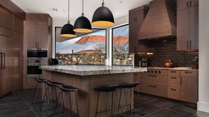 A modern kitchen with dark wood cabinets, a marble island, black bar stools, and a large window with a mountain view.