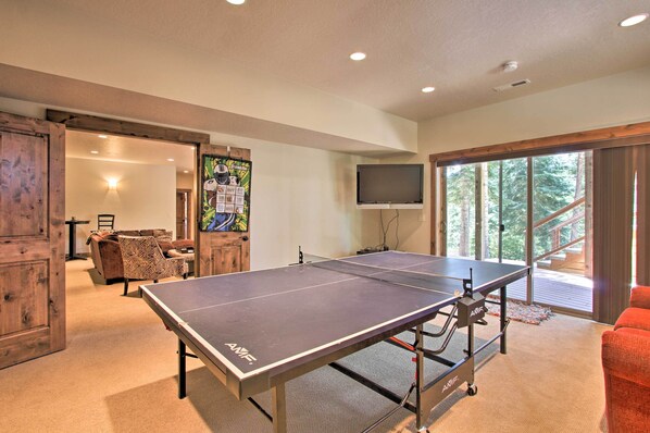 Una mesa de ping pong en una habitación con un ventanal grande, un televisor y una puerta de madera.