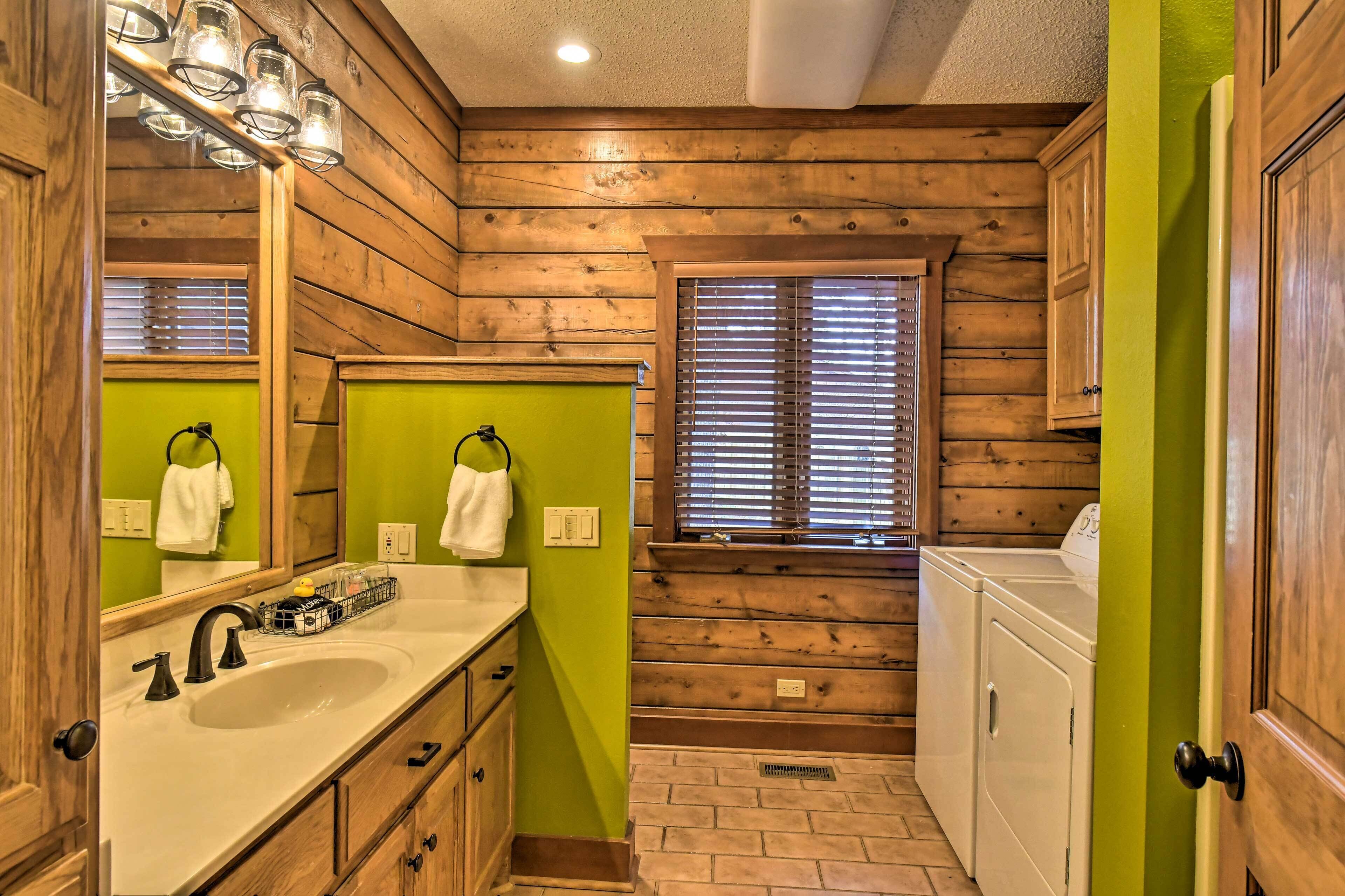 Une salle de bain avec des murs en bois, un mur d’accent vert, un lavabo avec un robinet noir et une fenêtre avec des stores.