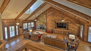 A spacious loft-style living area with a stone fireplace, wooden beams, and a large TV. - Hot Tub, Grill: Hilltop Log Cabin in Hot Springs! (Hot Springs)