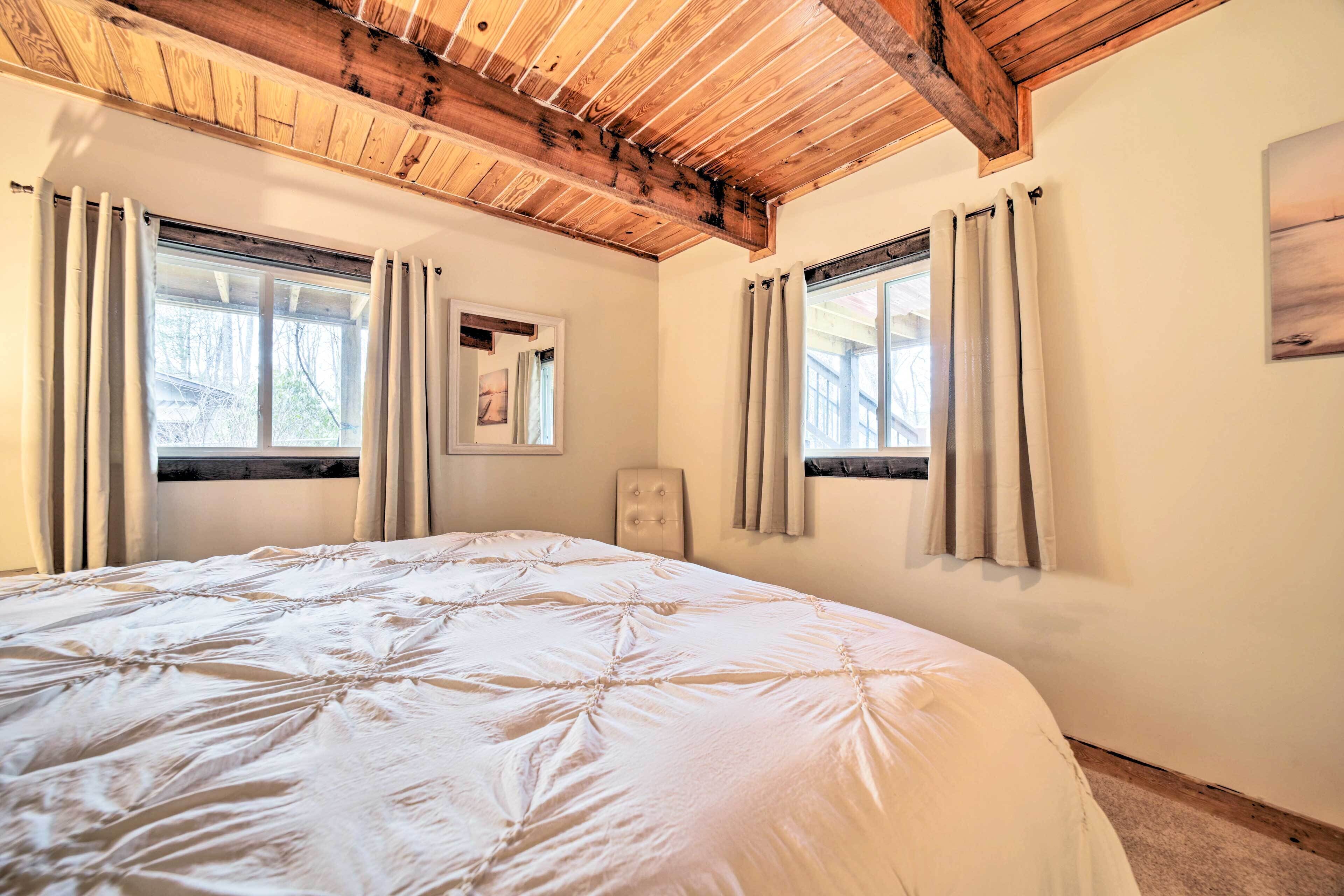 A bedroom with a large bed, wooden ceiling beams, two windows with curtains, and a framed picture on the wall.