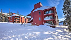 Un immeuble rouge à plusieurs étages, doté de balcons et d’un toit enneigé, entouré d’arbres et surplombant un ciel bleu clair.