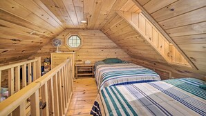 A cozy attic bedroom with wooden walls and a sloped ceiling. There are two beds with striped bedding, a small desk with a chair, and a window providing natural light. - Riverfront Lewiston Cabin w/ Stone Fireplace! (Mio)