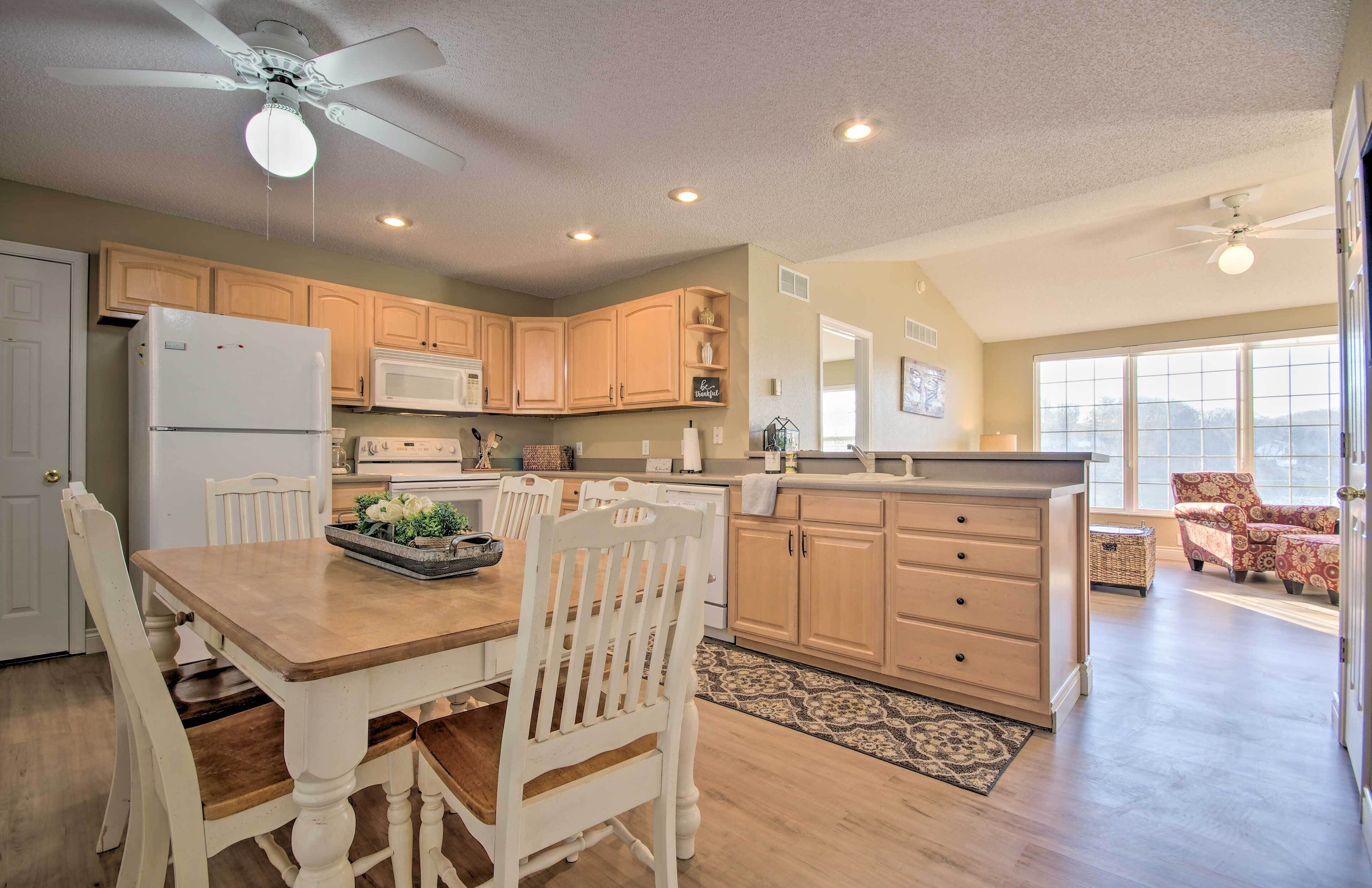 A spacious kitchen with wooden cabinets, a dining table, and chairs. There is a ceiling fan, recessed lighting, and a view of the living area through large windows.