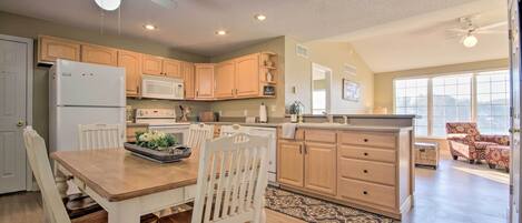 A spacious kitchen with wooden cabinets, a dining table, and chairs. There is a ceiling fan, recessed lighting, and a view of the living area through large windows.