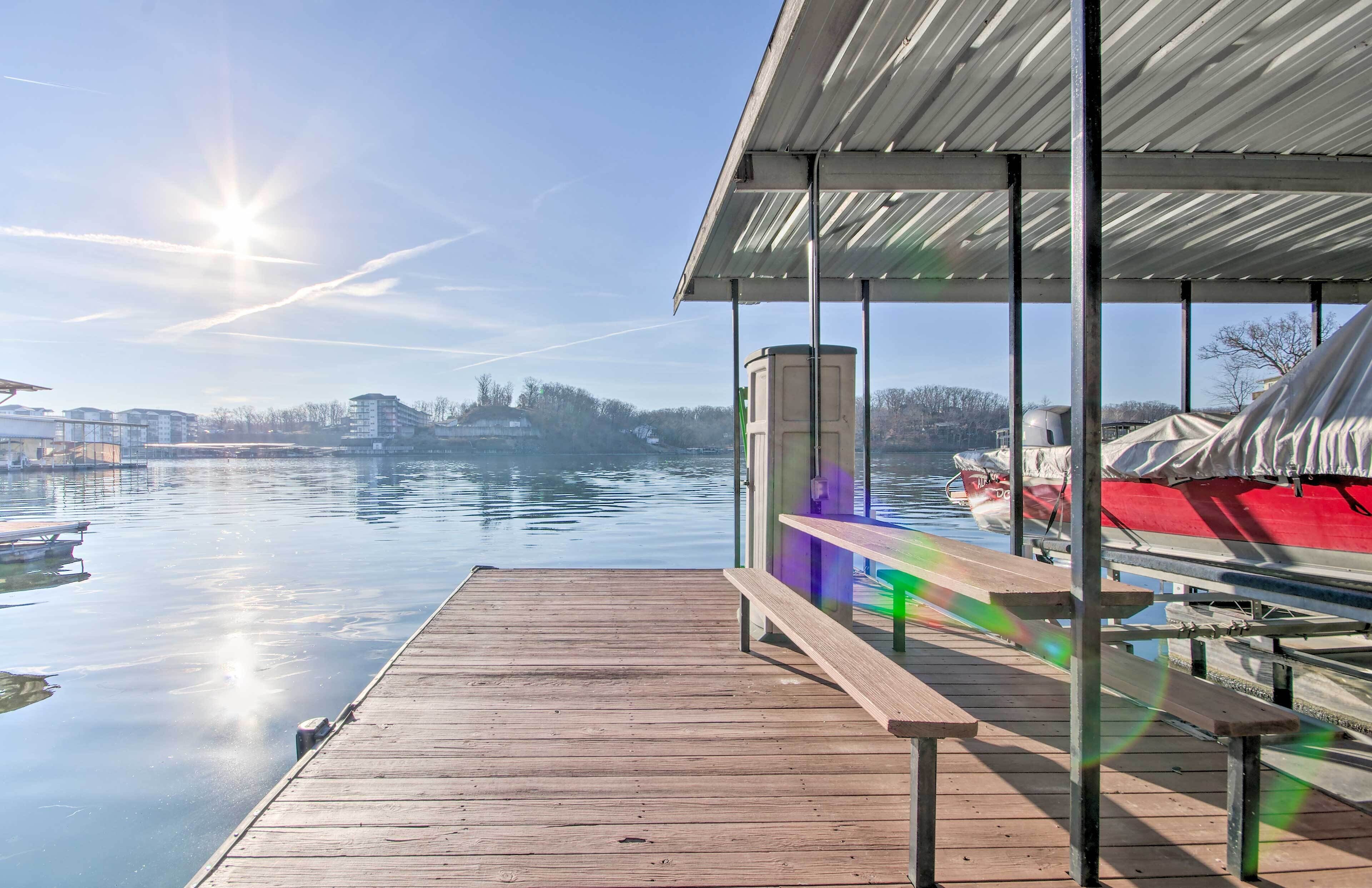 A wooden dock extending over calm water, with a covered seating area and a view of boats and buildings in the distance.