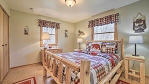 A cozy bedroom with a wooden bed frame, plaid bedding, and a small bedside table with a lamp. - Family Cottage 3 Mi to Higgins Lake State Pk! (Roscommon)