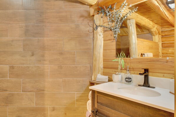 Une salle de bain rustique, avec un sol en bois, un meuble-lavabo en bois doté d’un évier, un miroir et un porte-serviettes.