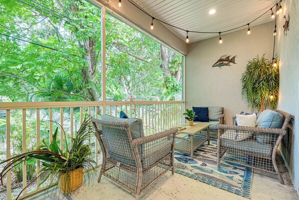 A cozy outdoor seating area with wicker furniture, a table, and a large window overlooking lush greenery.