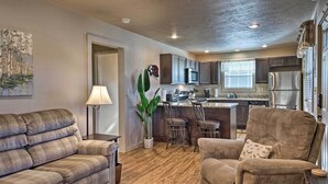 A living room with a sofa, armchair, and a coffee table. There is a kitchen in the background with a refrigerator, microwave, and cabinets. - Charming Cabin w/ Patio - Walk to Table Rock Lake! (Lampe)