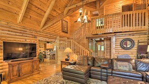 Interior espacioso de una cabaña de madera con una televisión grande, sofás de cuero, una escalera y techo de madera.