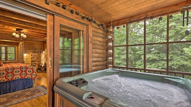 Interior de una cabaña de troncos con un jacuzzi, pisos de madera, una cama con colcha estampada y vistas al bosque a través de amplias ventanas.