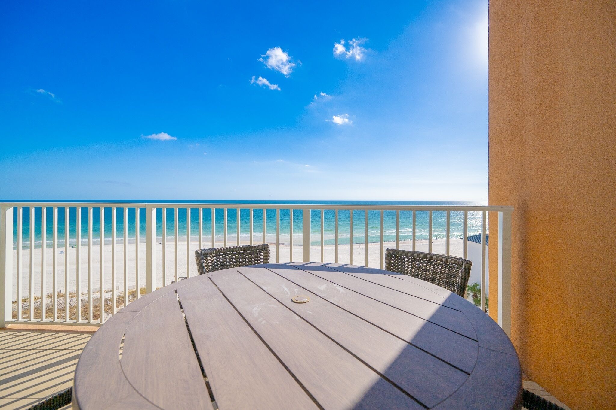 A round wooden table with two wicker chairs on a balcony overlooking a beach.