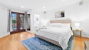 A bedroom with a large bed, wooden floor, a blue patterned rug, and a door leading outside.