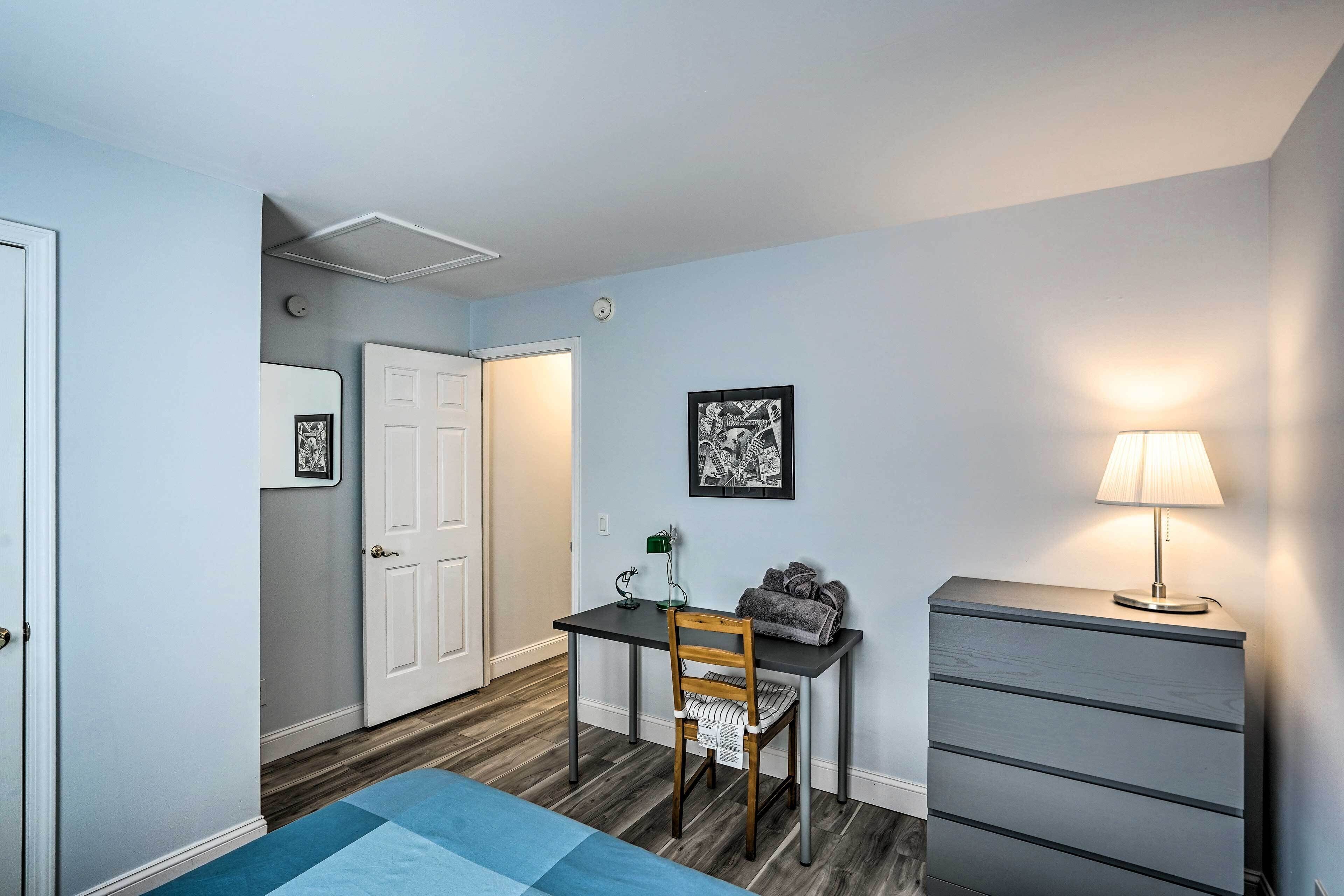 Une chambre à coucher de taille modeste, comprenant un lit, un bureau avec une chaise, une commode, une lampe et une porte.