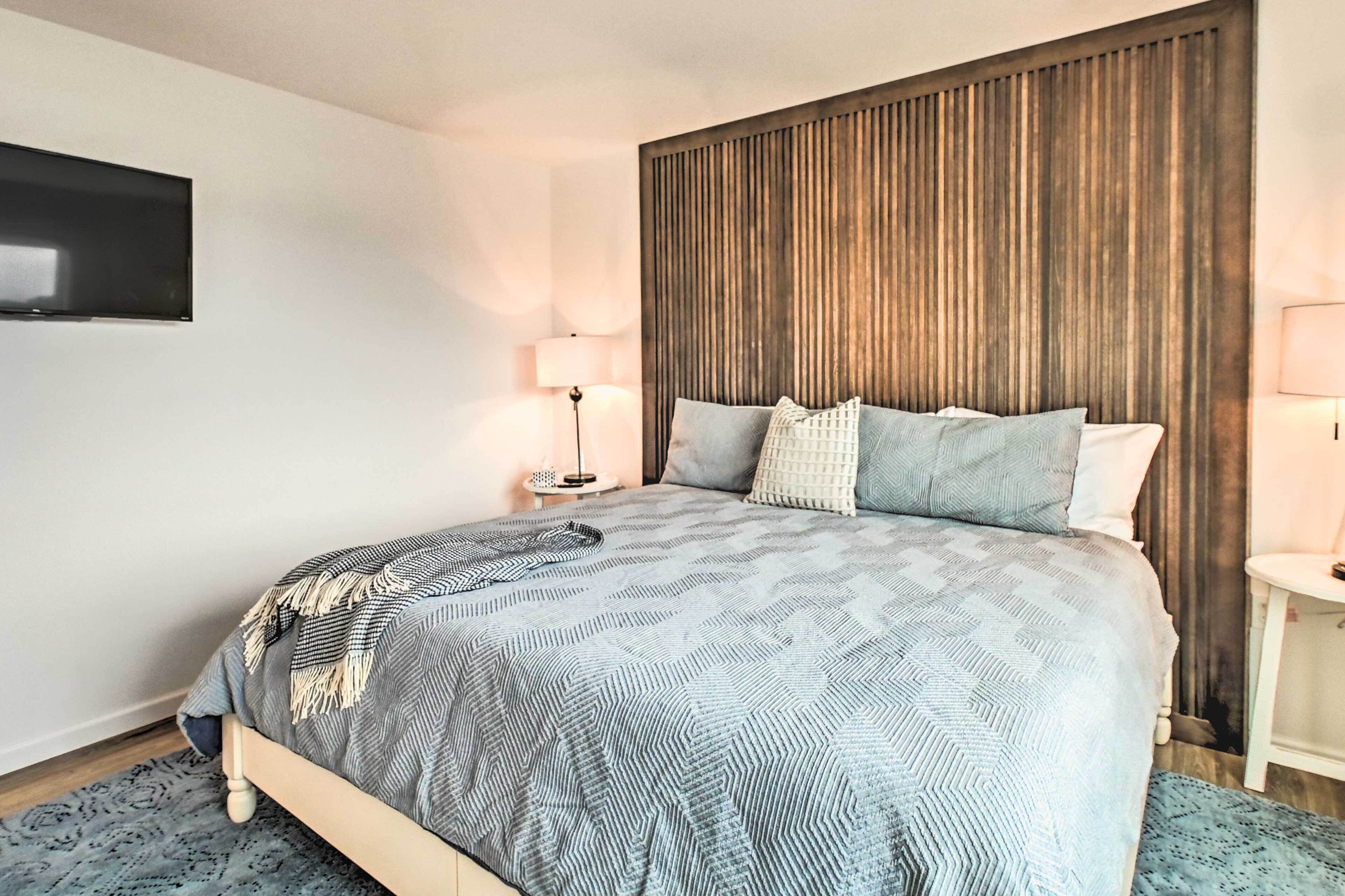 Een moderne slaapkamer met een groot bed, een houten hoofdbord, twee nachtkastjes met lampen en een flatscreen televisie aan de muur.