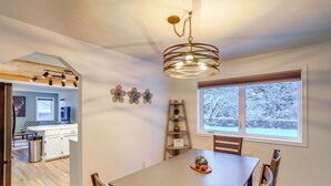 Un coin repas avec une table, des chaises, un lustre, une fenêtre donnant sur des arbres et, au fond, une cuisine.